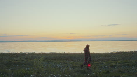 woman holding signal lantern, walking along scenic lakeside at sunset