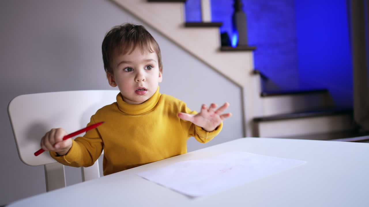 Small toddler in yellow sweater sitting at desk. Kid holds a red pen and waving it. Child drawing at home.