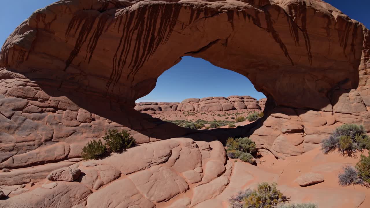 Inquadratura grandangolare che cattura un arco di roccia naturale sotto un cielo azzurro limpido, enfatizzando la vastità.