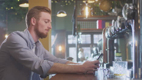 hombre infeliz sentado en un pub bebiendo solo con un vaso de whisky enviando un mensaje en el teléfono móvil