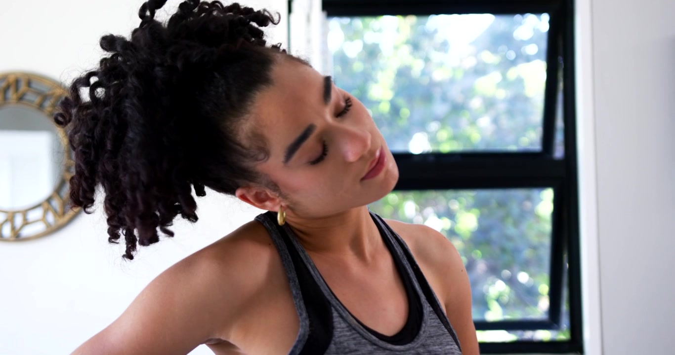 Woman starting closed eyes, standing by window, doing neck rolls for warmup in tank and watch
