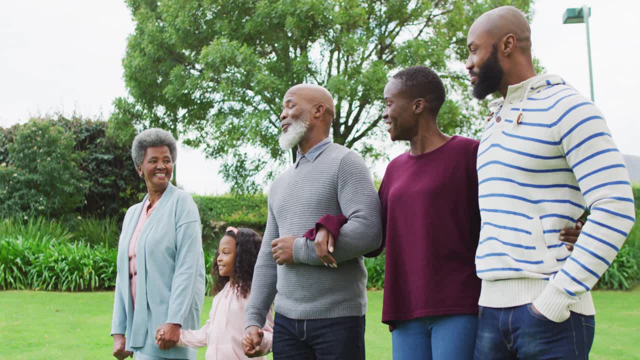 Video of happy african american parents and grandparents walking with granddaughter in garden