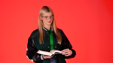 Pretty-Blonde-Student-Standing-Reading-a-Book-Against-Red-Background,-Studio-Shot