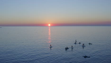 Grupo-De-Paddle-Boarders-En-El-Agua-Al-Atardecer-Viendo-La-Puesta-De-Sol-Bajo-El-Horizonte