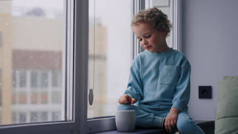 charming little boy is looking out window sitting on still watching falling snow in winter day