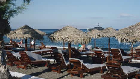 people at oceanside beach lounge relaxing under wooden umbrellas in chairs near water relaxing chairs at beach lounge overlooking oceanside with mega yacht near in santorini, greece