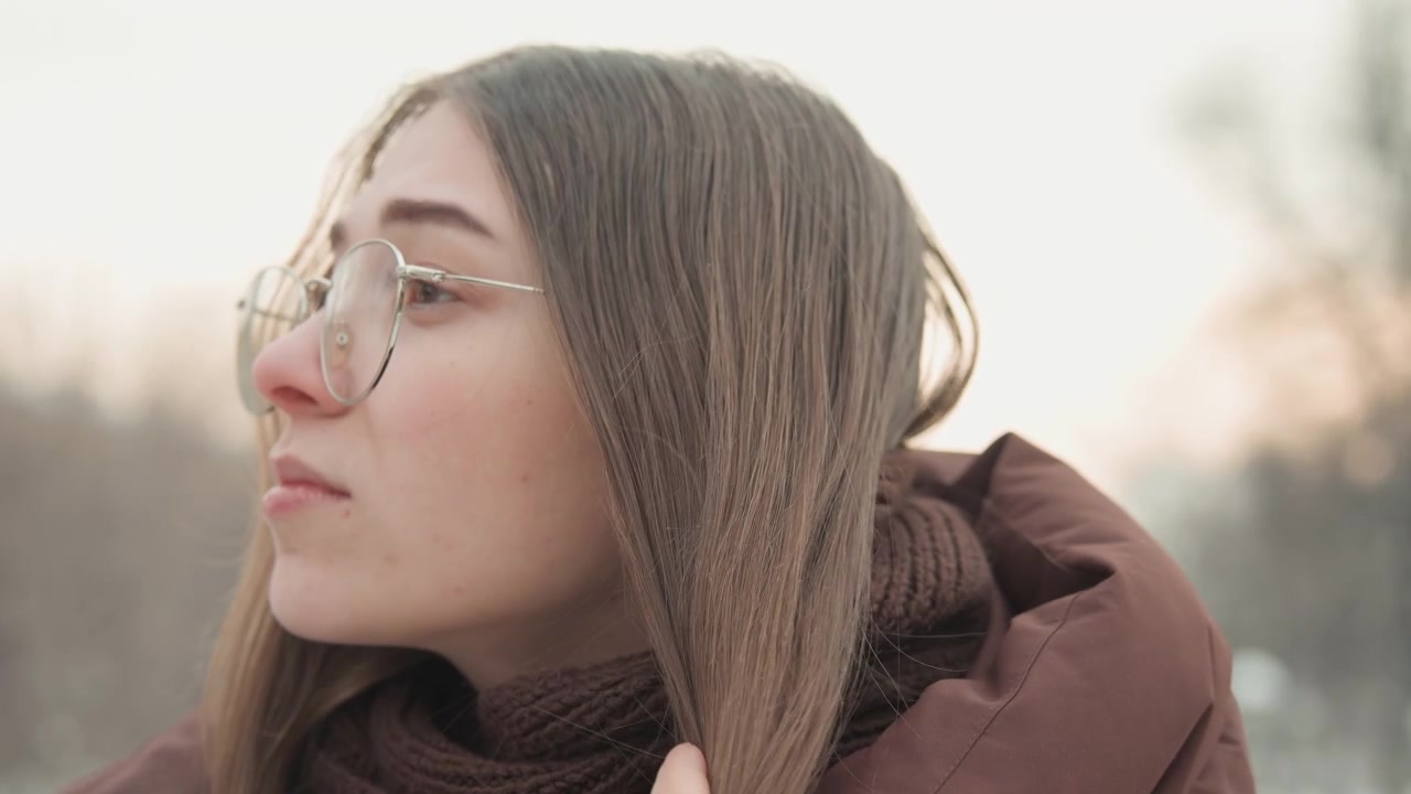 Profilo a tre quarti sinistro di una giovane donna che controlla capelli e cuoio capelluto separando le ciocche e pettinando le radici, con scaglie bianche visibili che suggeriscono cuoio capelluto secco o residui di prodotto in una luce naturale soffusa.