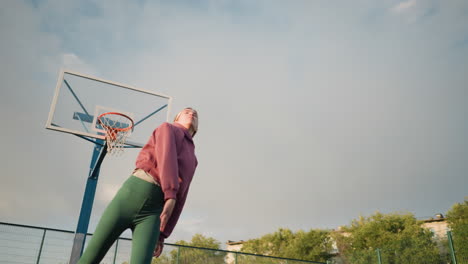 lady in athletic wear jumps high, slamming ball towards hoop on outdoor court, basketball hoop visible in background, surrounded by greenery, fence, and buildings under warm sunlight