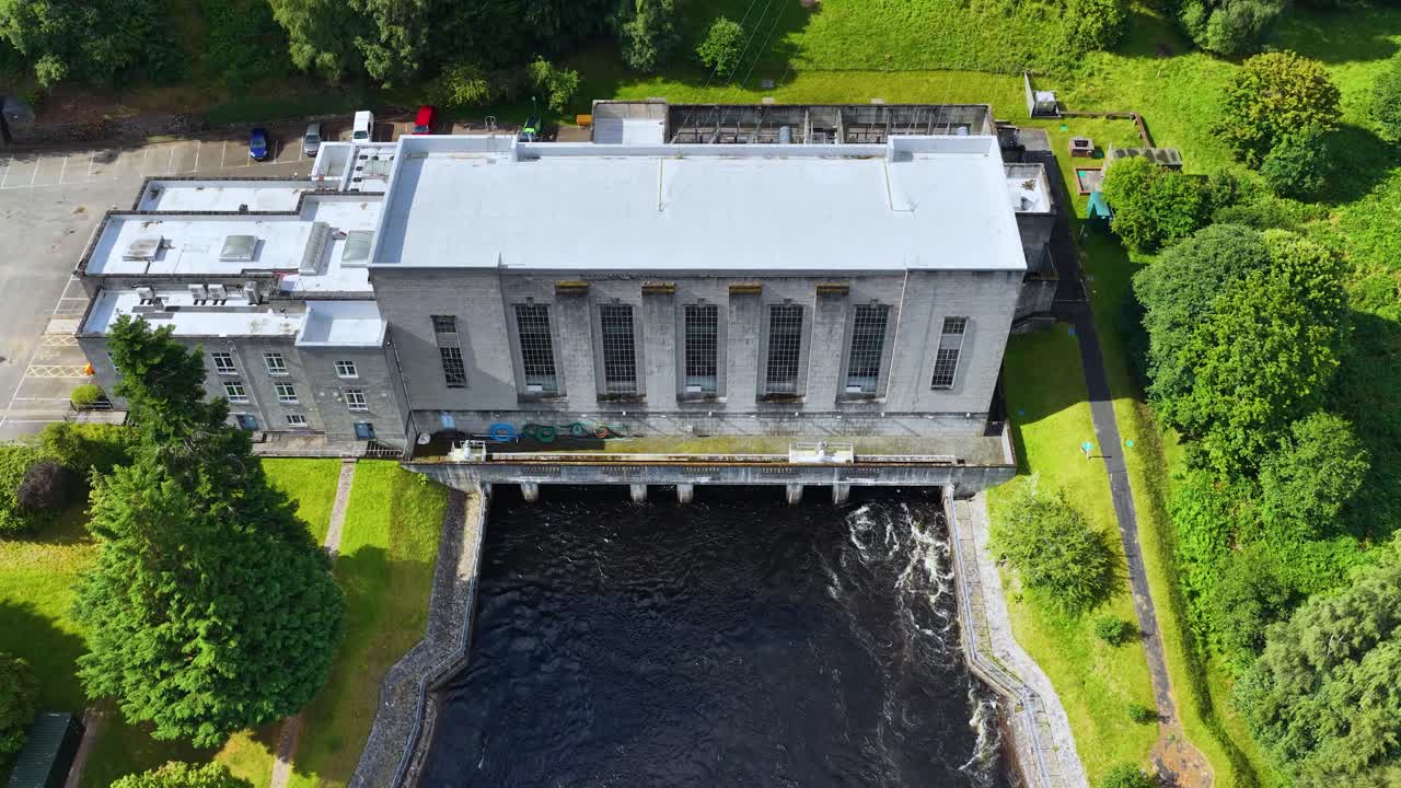 Drone captures hydroelectric power station, river, and green landscape in bright daylight, overhead perspective