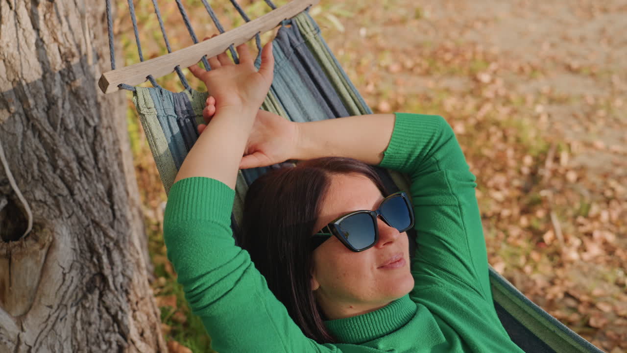 Donna che si rilassa in amaca in campeggio, con le braccia alzate sulla barra distanziatrice, occhiali da sole e maglione verde. Fogliame autunnale e luce soffusa, in un sogno ad occhi aperti contemplativo e una pausa consapevole, una viaggiatrice solitaria che si concede un momento.