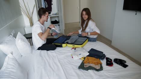 stylish young couple sitting on bed with opened suitcase and packing up things for travel. man and woman thinking about needed