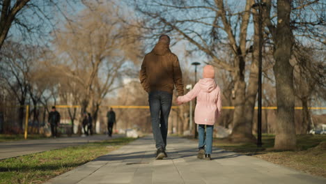 father in a brown jacket walks hand-in-hand with his little daughter wearing a pink cap and jacket, through a park