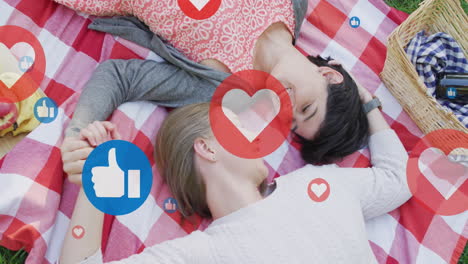 animation of hearts and like icons over caucasian female couple having picnic