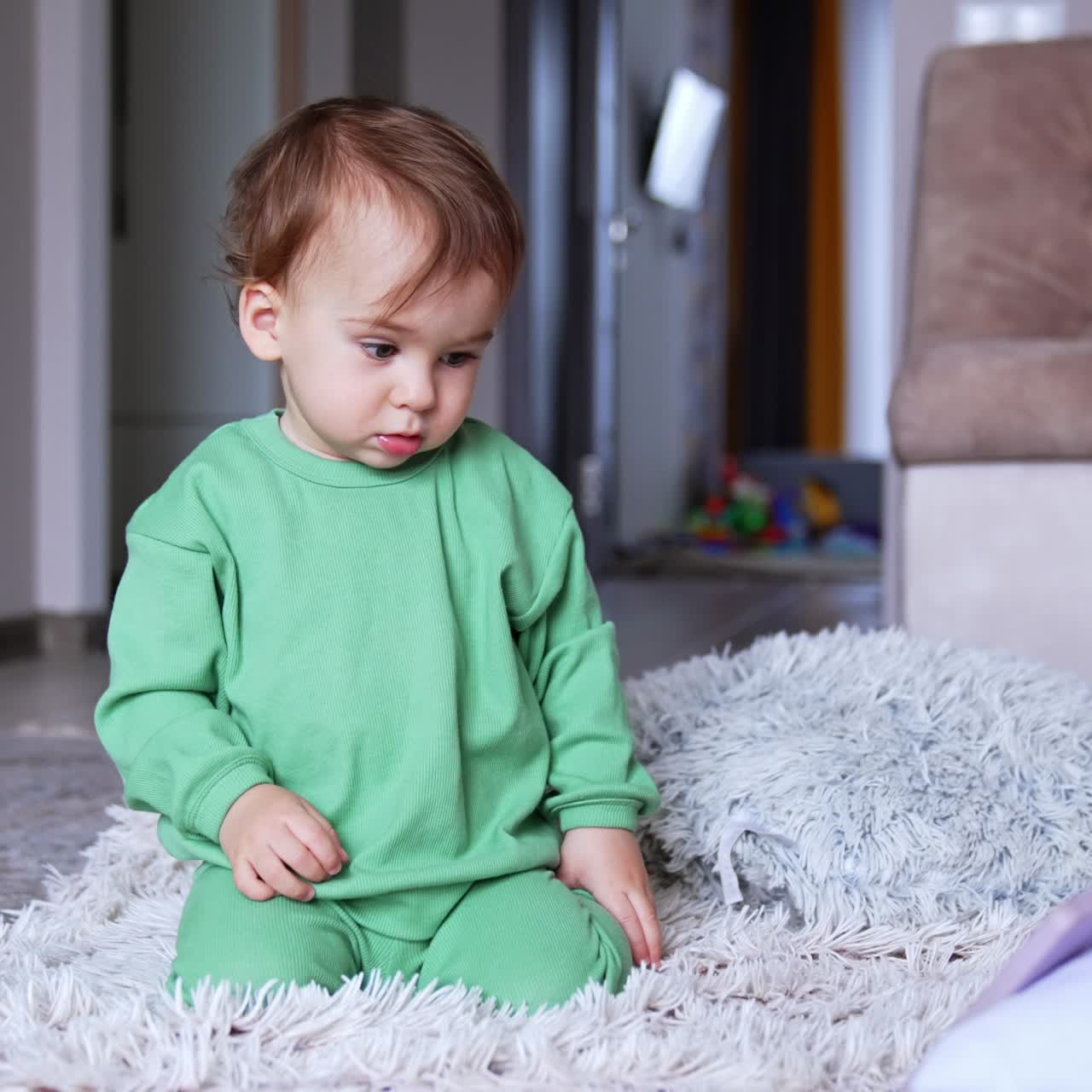 Lovely calm boy of one year old sits on a soft carpet in the room. Kid in light green sport suit watching videos on the phone in front of him