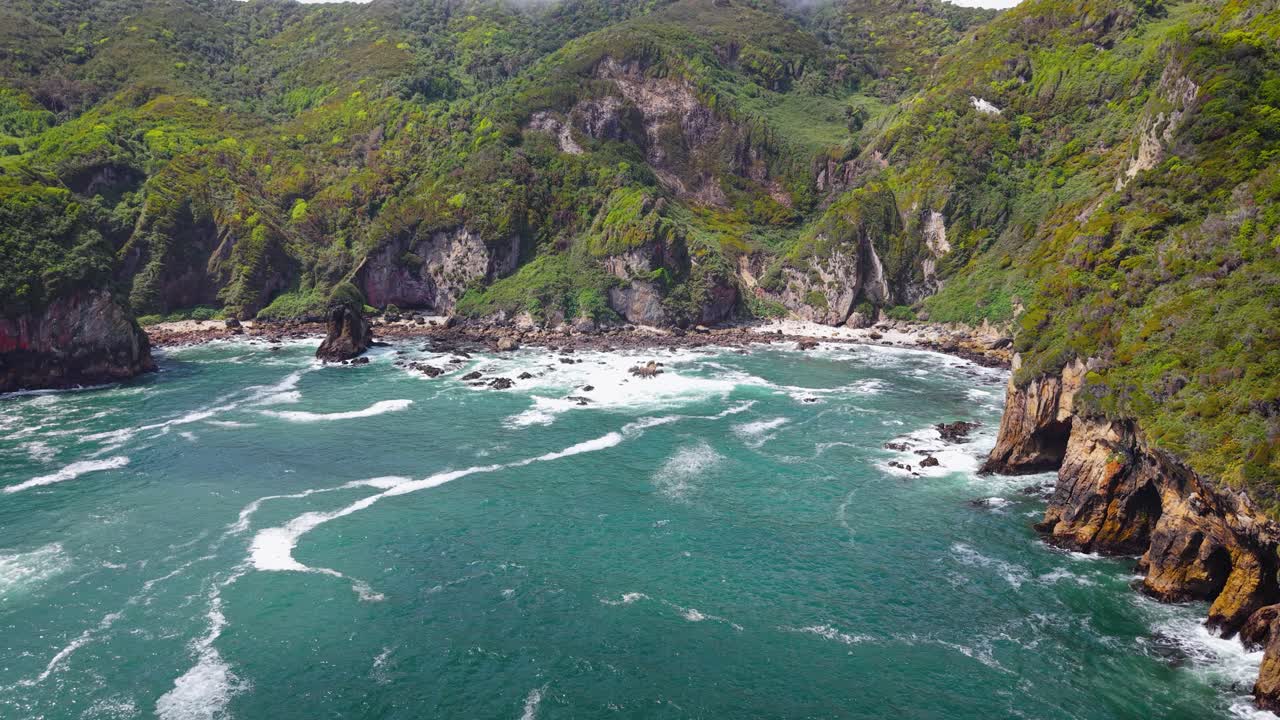 Vista aérea de drone de um monte de pedras coberto de grama com uma gruta marinha na ilha de Chiloé, costa do Pacífico da Patagônia, Chile. Ondas fortes atingem uma praia arenosa remota e penhascos verdes e acidentados sob um céu nublado