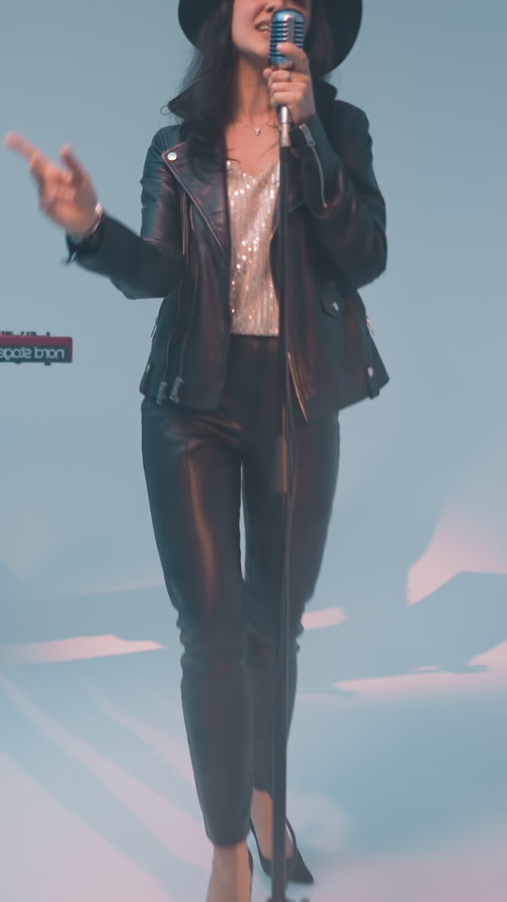 A vocalist in a black leather jacket, trousers, and hat stands at a vintage microphone held with one hand. A keyboardist plays a red sampler against a blue background as the camera zooms in and out