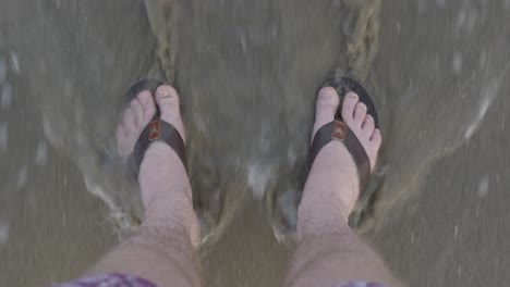 men feet in sandals stand on the beach - waves come easily to the beach and cover the feet