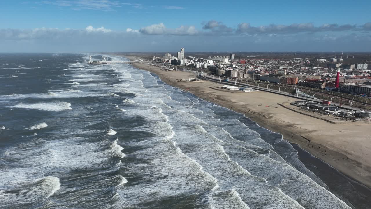 amplio camión aéreo dejó una toma de muchas líneas de olas de una tormenta que se estrellaba en la playa con una ciudad, un muelle y una rueda de ferris en el fondo