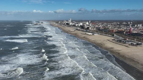 wide aerial truck left shot of many lines of waves from a storm crashing on the beach with a city, pier and ferris wheel in the background