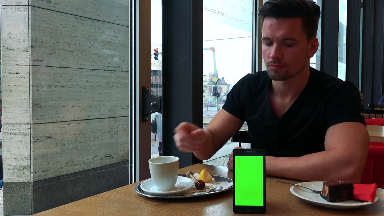 un joven apuesto se sienta en una mesa en un café, come pastel y bebe té, un teléfono inteligente con una pantalla verde en primer plano