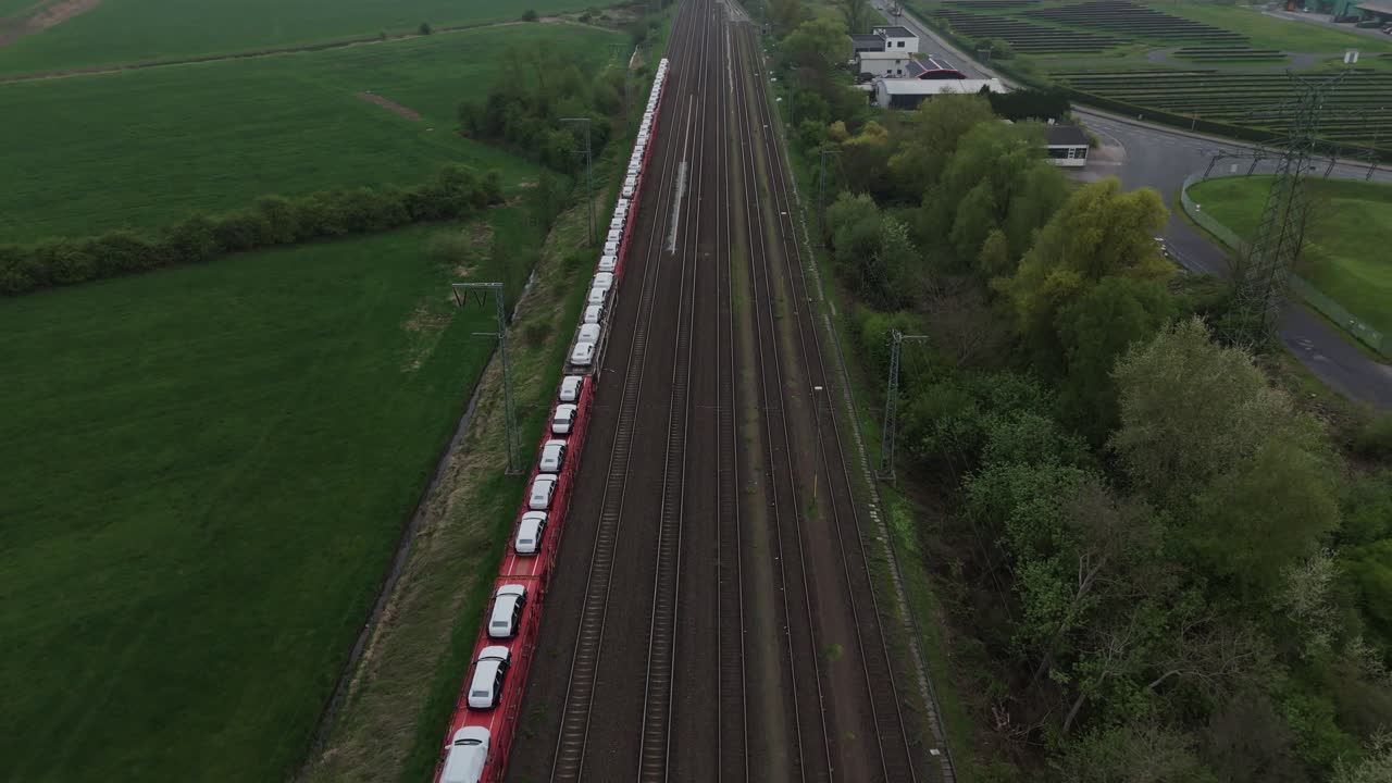 Aerial shot of railway tracks near Leer, Germany, on a cloudy afternoon. Cargo train with white vehicles on red flatcars. Wide landscape view with green fields and industrial surroundings. Shot in 4K.