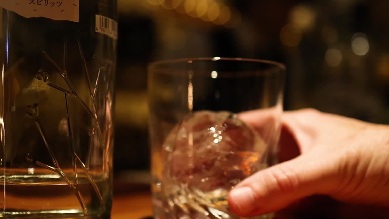 A hand gently holds a glass containing a large ice sphere, set against a dimly lit background.