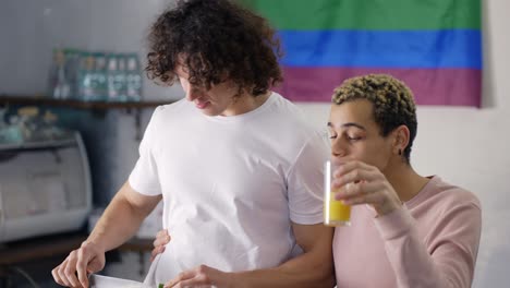 Hermosa-Pareja-Gay-En-La-Cocina-Juntos-Preparando-El-Desayuno,-Bandera-Lgbt-En-El-Fondo