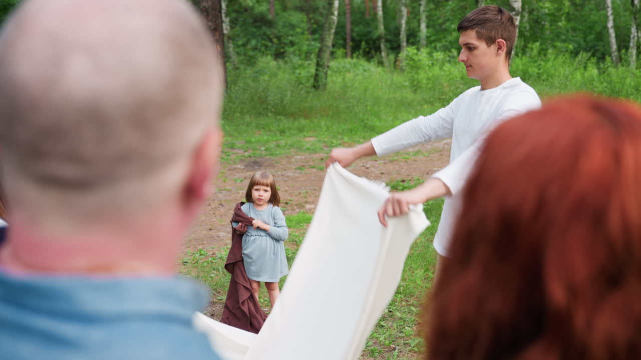 Вид сверху: родители смотрят, как дети расстилают ткань для пикника на траве, а младший ребенок держит коричневое одеяло в спокойной лесной обстановке. Солнечный свет, проникающий сквозь деревья, создает теплую семейную атмосферу любви.