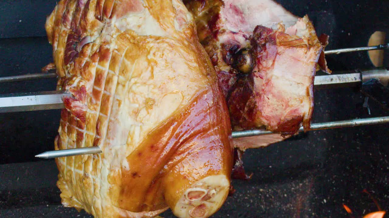 Juicy pork knuckle slowly rotates on spit above wood fire, outdoor market, natural lighting