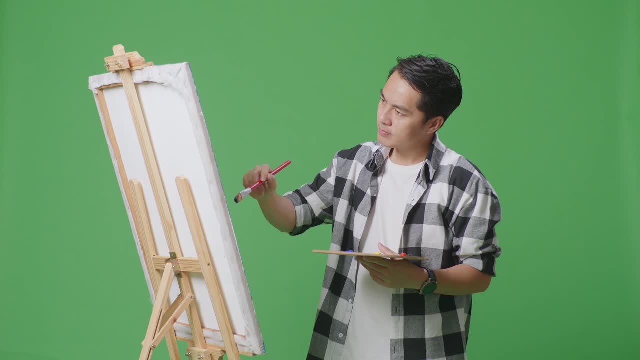 아시아 남성 예술가 는  을 들고 그림 개념 을 고민 하고 있으며, 그린 스크린 스튜디오 에서 오일 페인트 와 브러쉬 로 캔버스 에 그림 을 그리고 있다