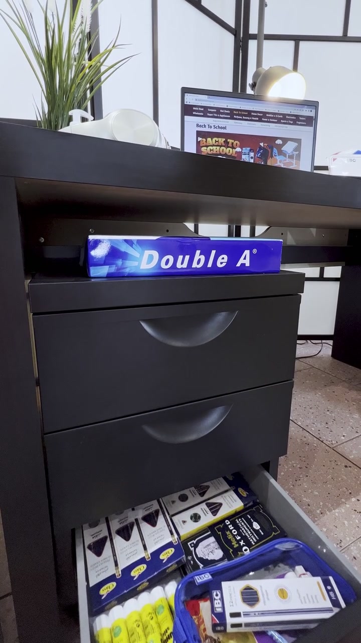 Hand opens a black office drawer filled with Double A paper, notebooks, pens, and a pencil case beneath a laptop displaying a back-to-school page in a bright indoor workspace.