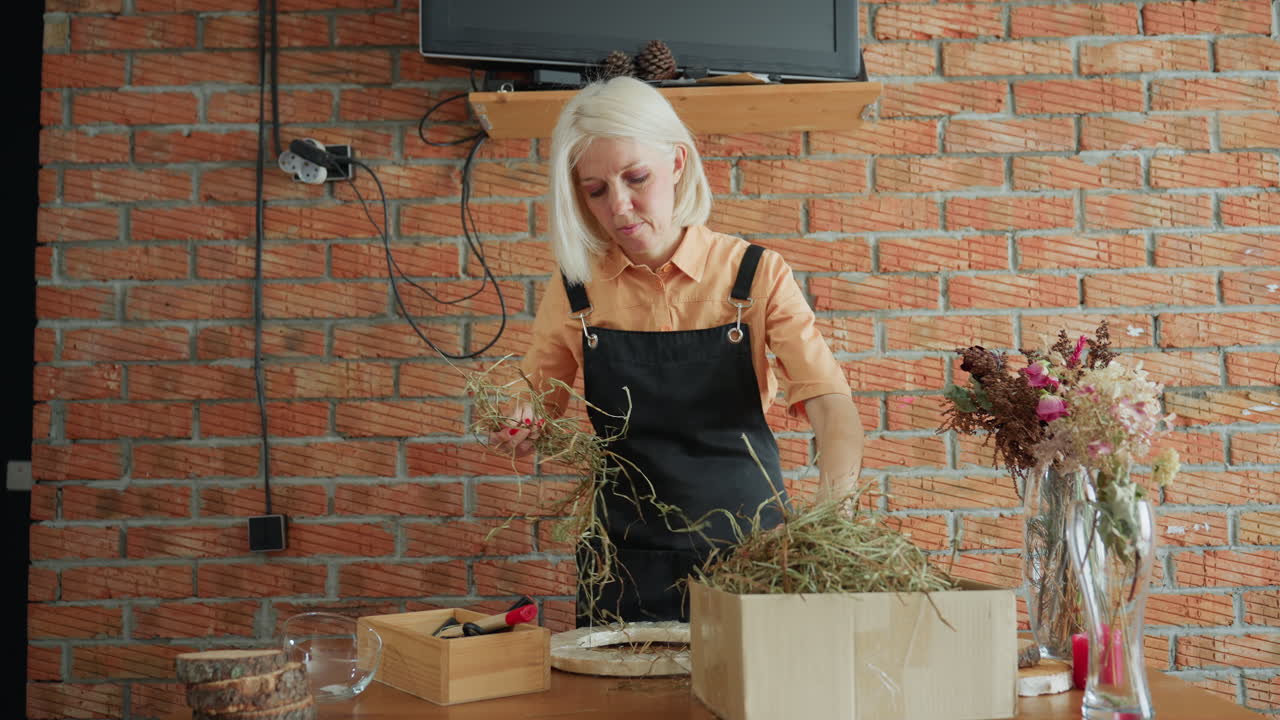 Florist in apron works with natural dried grass material at wooden table, preparing arrangement with craft supplies and rustic elements, surrounded by dried flowers in vases against brick wall interior