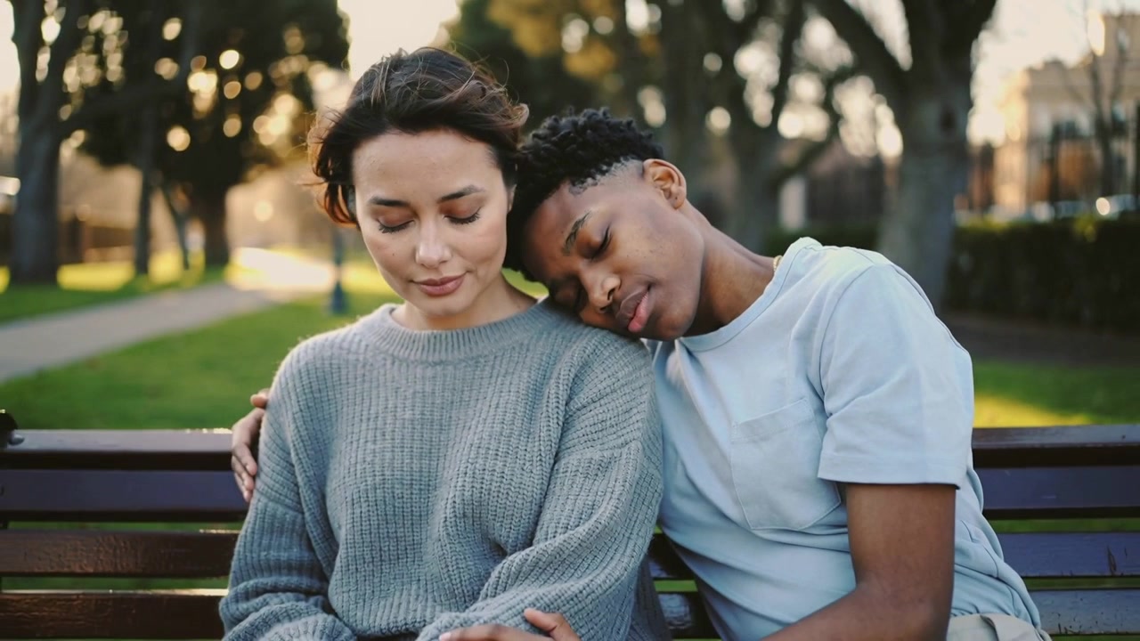 Leaning man resting head on partner's shoulder, wrapping arm for comfort on park bench
