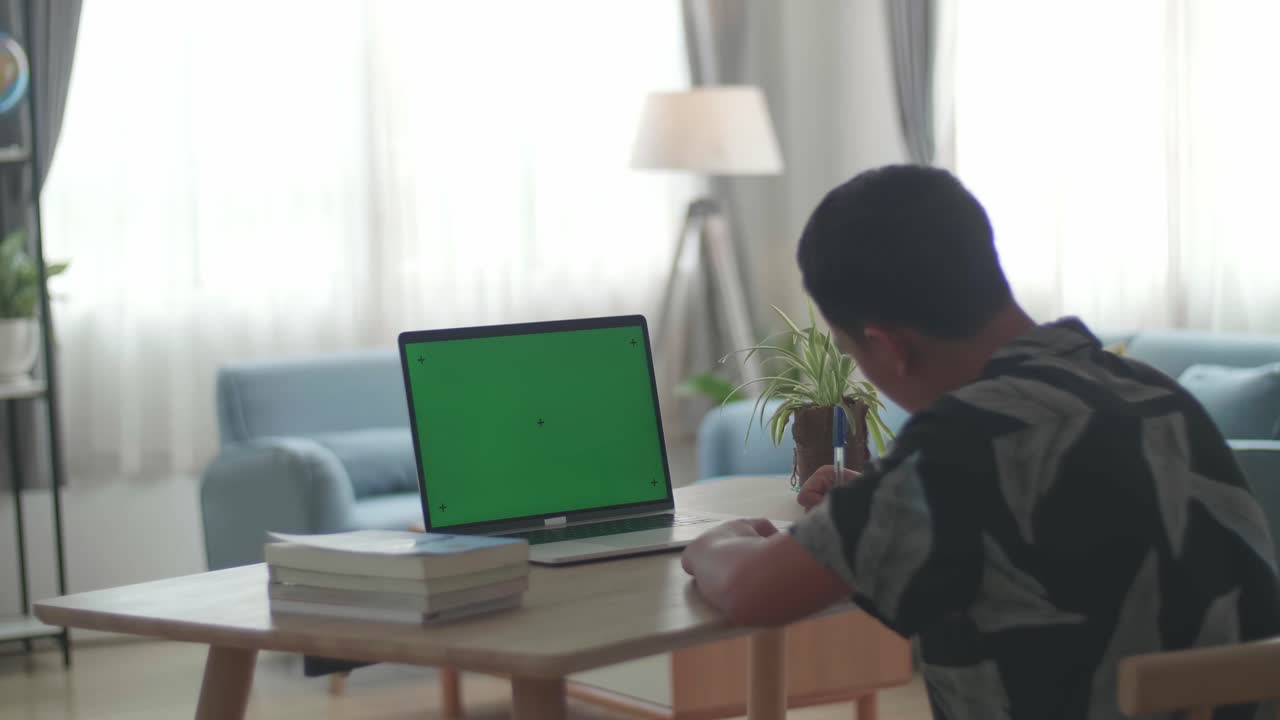 Teenage Boy Sitting At His Desk Learning Online On A Laptop With Green Mock-Up Screen At Home