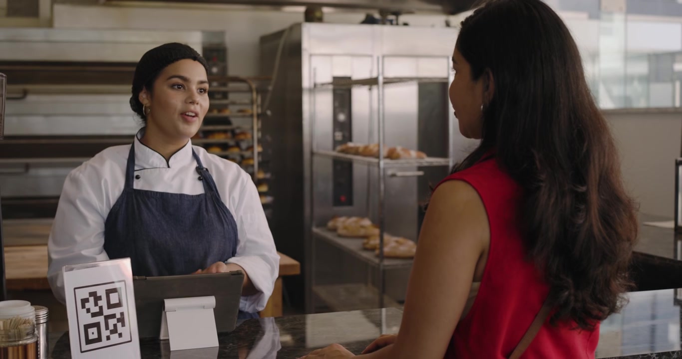 Female baker following female buyer request at counter taking bread in apron bagging, tapping card