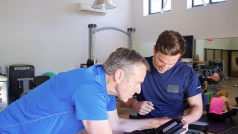 Senior-Man-Exercising-On-Cycling-Machine-Being-Encouraged-By-Personal-Trainer-In-Gym