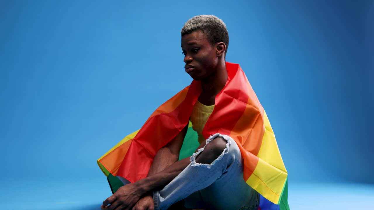 Giovane uomo nero seduto con bandiera arcobaleno su sfondo blu