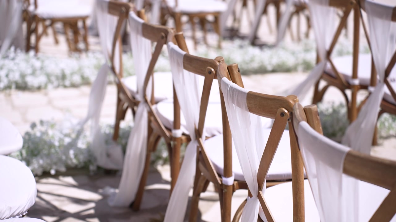 Sedie disposte in fila per un matrimonio. Tessuto bianco è legato su ogni sedia. L'area mostra alcune decorazioni floreali a terra. L'allestimento è pronto per l'arrivo degli ospiti.