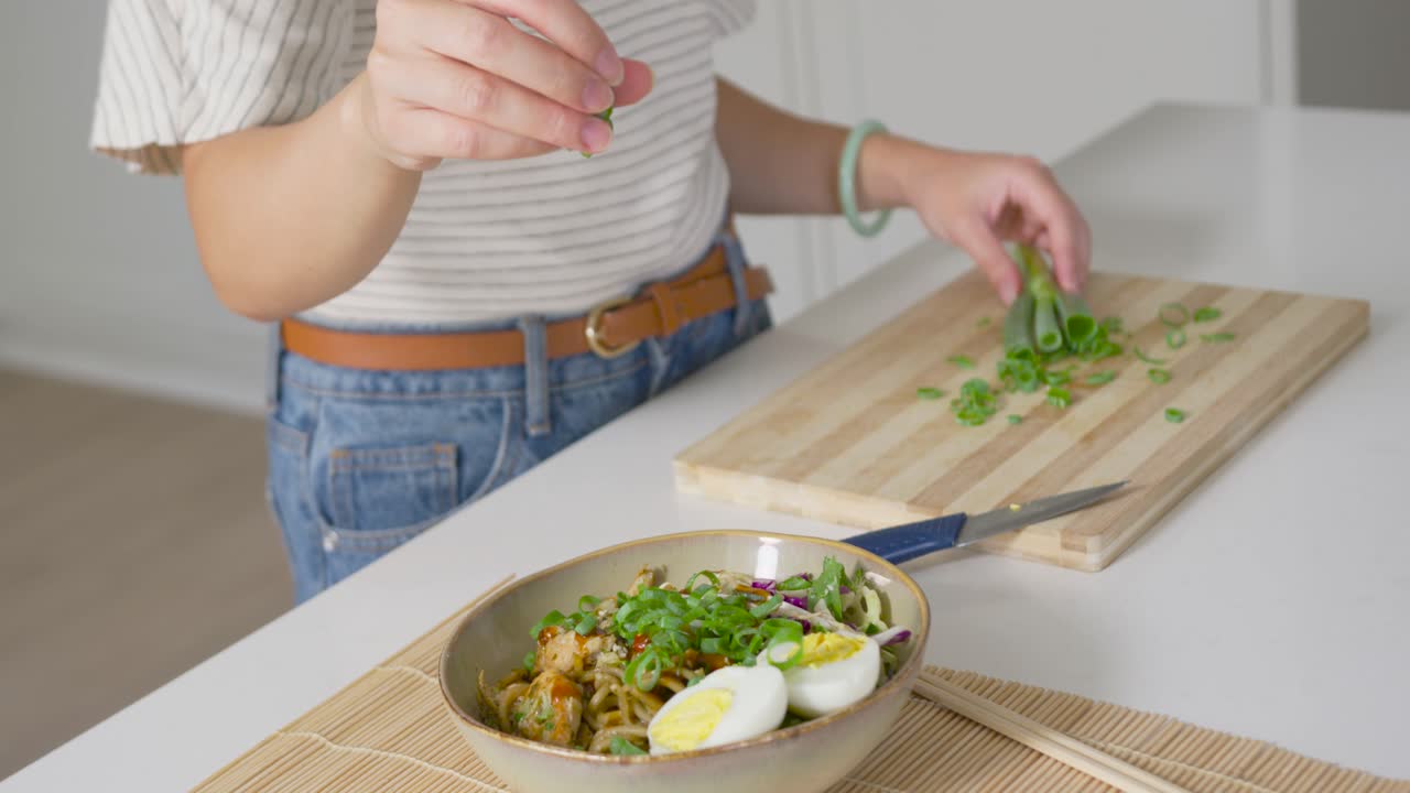 Mulher a guarnecer uma tigela de noodles na bancada de casa, a pegar cebolinhas da tábua de cortar para servir.