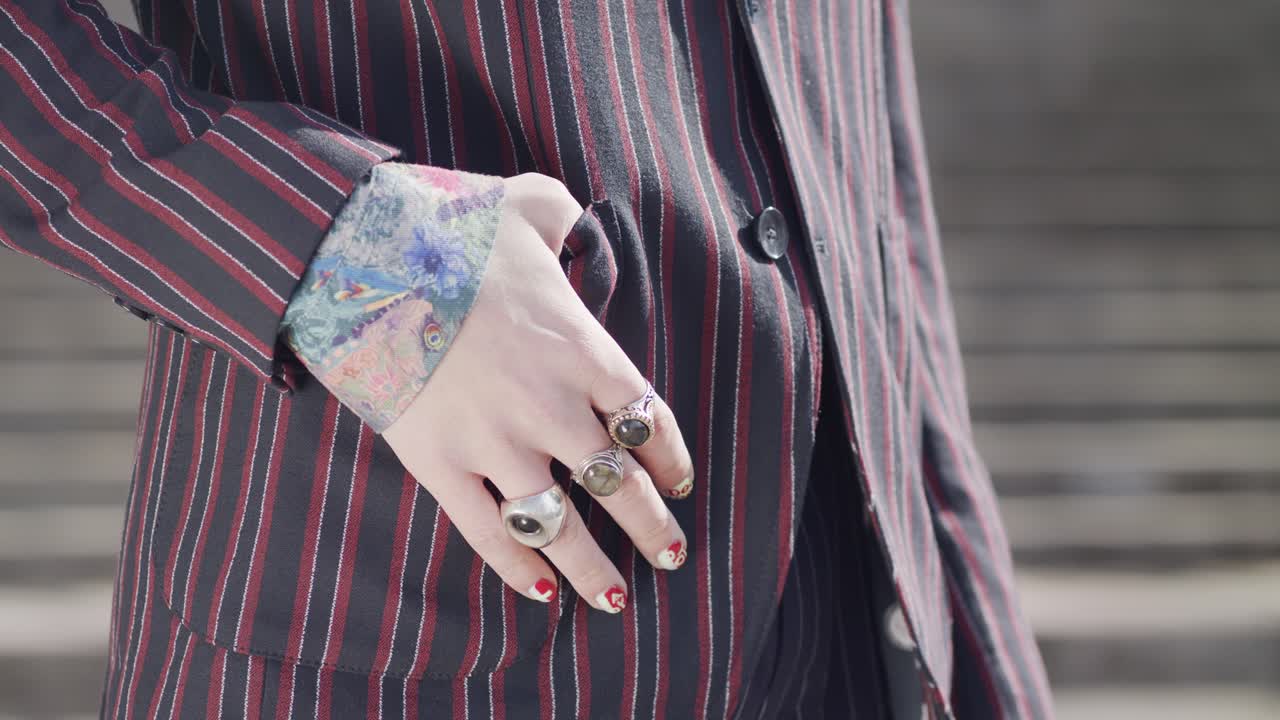A close-up on the stylish details of a person's outfit: a hand with large, ornate rings and painted nails rests against a pinstripe blazer with a colourful patterned cuff.