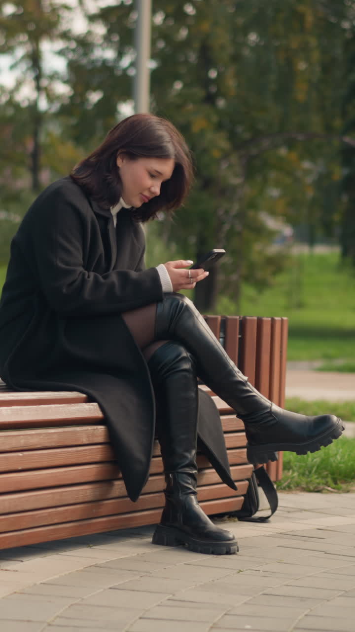 mujer sentada en un banco del parque usando un teléfono inteligente, vestida con abrigo y botas negras, rodeada de árboles verdes exuberantes, en un parque urbano con una vista borrosa de personas caminando en el fondo