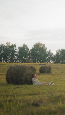 mujer sentada en el suelo, descansando contra una gran bala de heno en tierras de cultivo abiertas, paisaje rural pacífico con balas de heno esparcidas, luz cálida de la noche y un entorno rural sereno