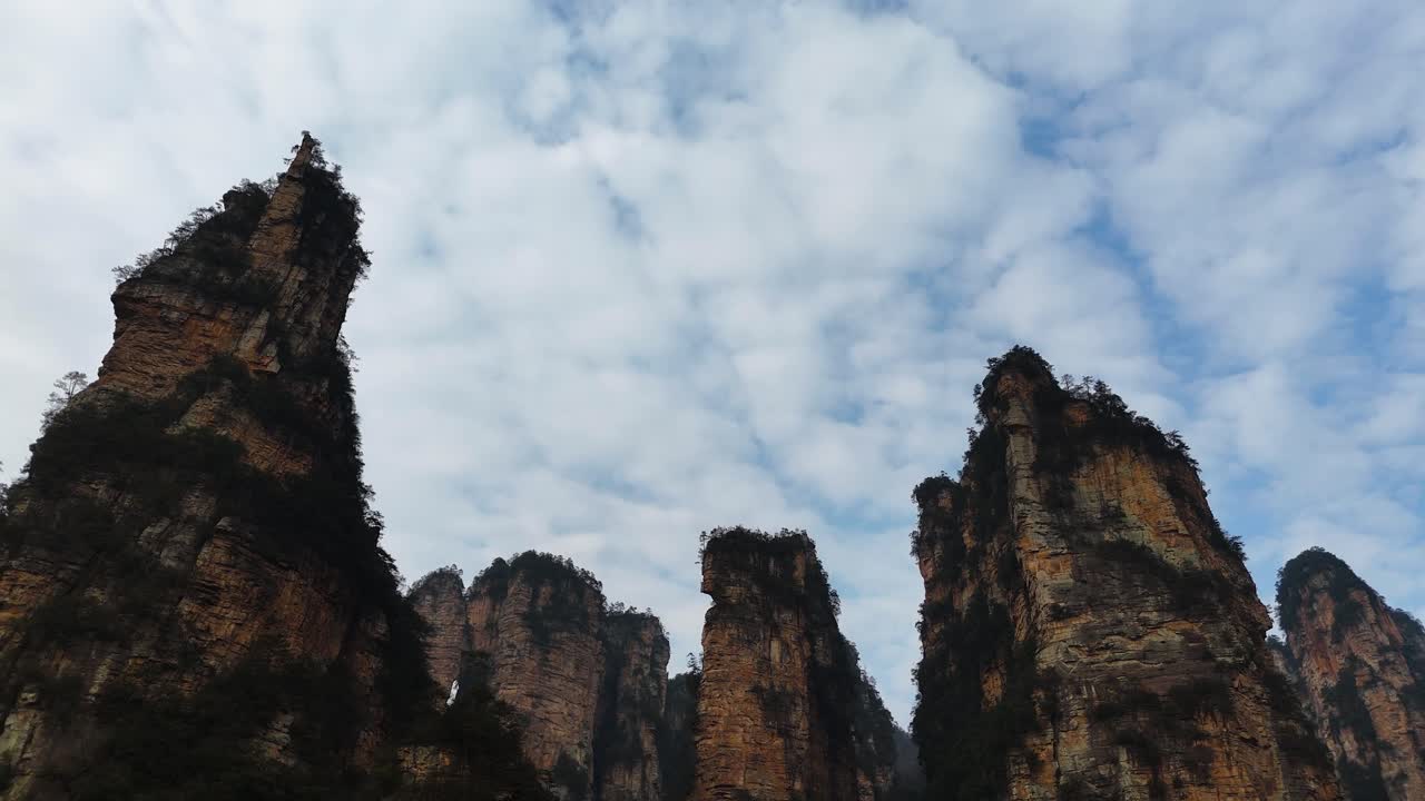 Towering sandstone formations reach towards the sky in Zhangjiajie, their rugged edges silhouetted against a backdrop of scattered clouds. A breathtaking view showcasing nature’s grandeur.