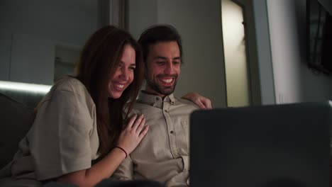 una feliz chica morena con una camiseta beige abraza a su alegre novio moreno con paja ven una comedia juntos en una computadora portátil gris por la noche en un apartamento estudio moderno