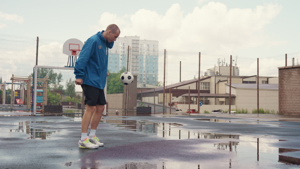 Allenamento di football solitario su campo bagnato, pozzanghere che riflettono lo skyline, uomo adulto in giacca blu che palleggia vicino al canestro da basket, lotto urbano vuoto sotto un cielo nuvoloso, palleggio costante e passaggio controllato
