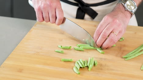 chef masculino cortando grandes trozos de judías verdes con su cuchillo en la tabla de cortar de su cocina