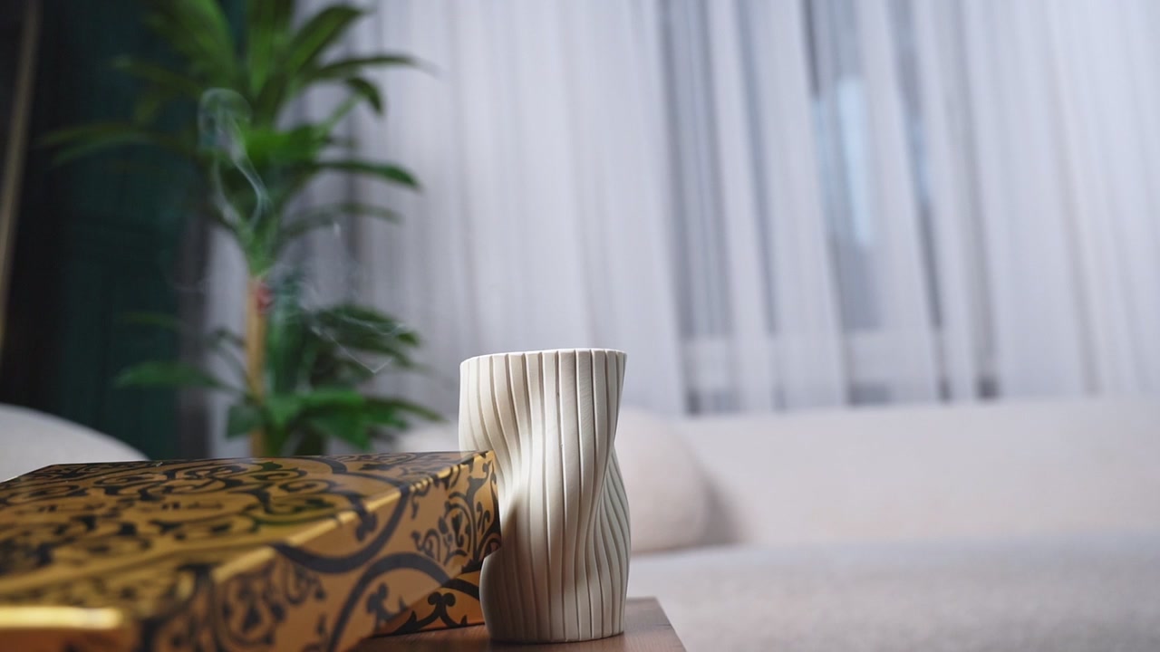 Gros plan sur la fumée d'encens qui s'élève à côté d'un vase en céramique blanc nervuré et d'une boîte à motifs dorés dans un salon minimaliste. La lumière douce du jour illumine l'environnement intérieur paisible de la maison.