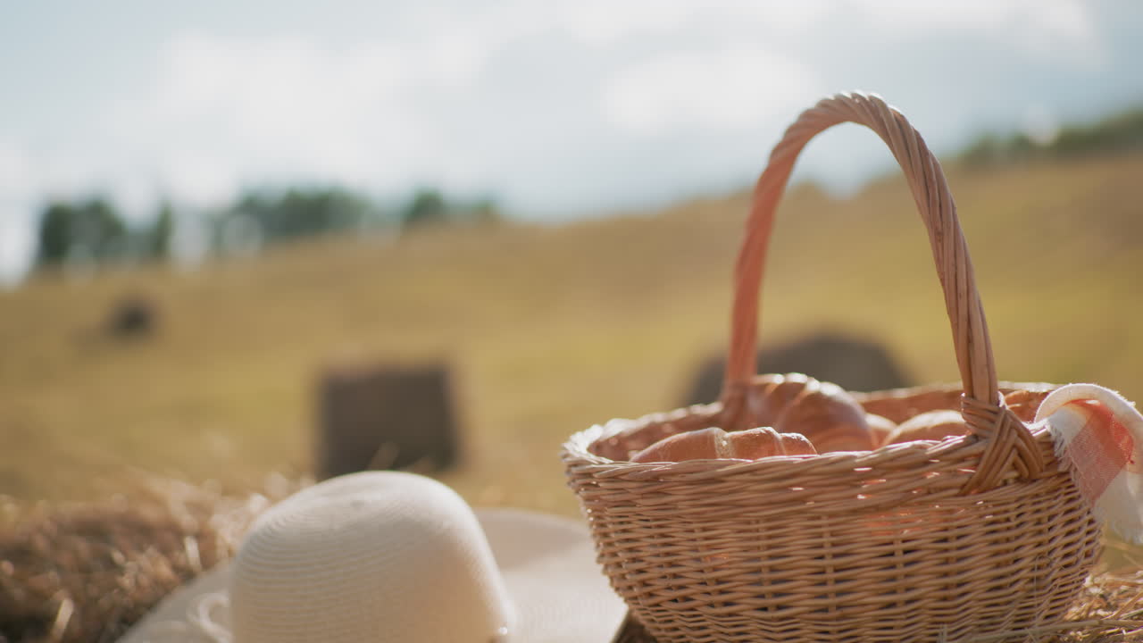 Slaměný klobouk a proutěný piknikový koš plný svačin položené na balíku sena v rozlehlé zemědělské krajině, malebná krajina s vlnícími se poli v pozadí evokuje klidnou venkovní pohodu a rustikální kouzlo.