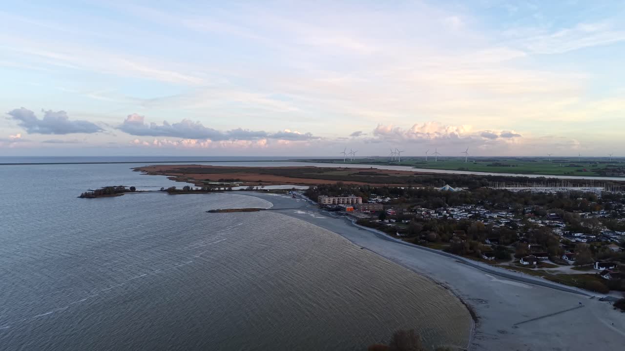 Aerial view of Dutch IJsselmeer coastline during ebb tide, showing shallow water patterns, wind turbines in distance. Calm shoreline under soft evening light in Netherlands. Wide shot