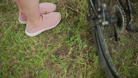 close-up of person s foot in pink sneaker removing bicycle stand, set on grassy ground with blurred background of bicycle wheel and lush green park